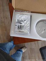 Opened box showing instruction manual and locking mechanism parts in plastic bag alongside white circular desktop stand base wrapped in plastic.