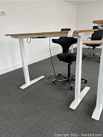 Side view of desk showing white steel legs, birch veneer top, and black office chair underneath.