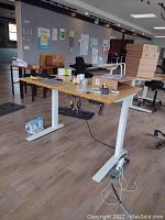 Full view of motorized adjustable height standing desk with bamboo top and white base, black ergonomic office chair visible behind in office setting.