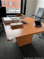 Square maple office table in an office setting next to chairs and windows. Items on table include electronic devices and boxes but are not included.