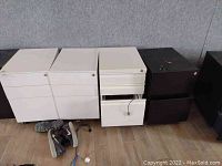 Four small office file cabinets, three black, one white, arranged on the floor with shoes nearby, one drawer open.
