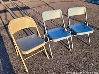 Three metal folding chairs outdoors in sunlight; two with blue padded seats and light gray backs, one beige metal chair.
