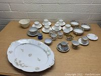 Various vintage china including dinner plates, tea cups with saucers, small bowls, and decorative cups with gold trim in different patterns arranged on a wooden table against a brick wall.