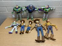 Front view of seven Buzz Lightyear and Woody toys arranged on wooden table, against brick wall background showing details and size differences.
