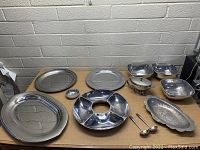 Assorted large and medium metal serving trays, divided serving platter, small serving bowls, sauce boat, and serving ladles arranged on a table.