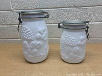 Both vintage milk glass canisters side by side, showing different heights and embossed floral and grapevine pattern on white opaque milk glass.