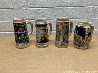 Four German beer steins arranged side by side showing their front decorative sides and handles, placed on beige surface against white brick wall background.