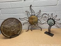 Three sun-themed decor items displayed against a brick wall: a round decorative plate with star and sun face design on left, a large metal sun sculpture with wavy rays and face in center, and a smaller sun sculpture with mirror center on right.