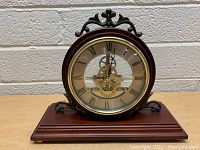 Full view of the mantle clock showing wooden base, glass face with Roman numerals, and visible brass gears inside.