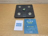 Top-down view of black Renpho smart scale with four silver sensors, two AAA batteries on top, instruction manual, and invitation card on beige wooden surface.