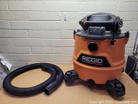 Front and side view of Ridgid wet/dry vacuum and detached black hose on a wooden surface against a brick wall.