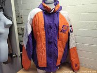 Front right angled view of the jacket displayed on a mannequin, showing the color blocking of purple, orange, and quilted white sections and the Phoenix Suns logo embroidered on the chest.