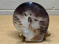 Front view of the hand-carved mother of pearl shell on the gold-colored stand showing detailed carving of a person walking near water and rocks.