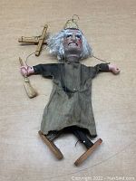 Full view of the marionette doll face-up on a table, showing its worn garment, wooden shoes, and broom accessory.
