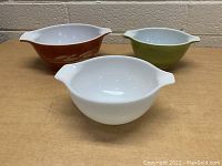 Three vintage Pyrex mixing bowls set on a wooden surface against a cinder block wall. One bowl is solid white, one is olive green with white interior, and one is brown with cream leaf pattern.