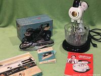 Full lot photo showing the Sunbeam Mixmaster with glass mixing bowl and beaters, boxed General Electric iron, Westinghouse thermometer set in original packaging, and instruction manuals.