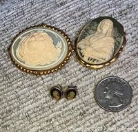 Three cameo jewelry pieces displayed with a quarter for size reference: two large oval pins and one pair of small stud earrings. Background is carpeted.