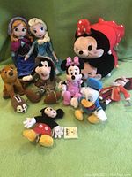 Full view of all eleven Disney plush toys arranged on a green background including Frozen characters, Mickey and Minnie Mouse, Donald Duck, Goofy, Chip and Dale, and Mad Hatter.