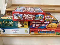Five board game boxes stacked on stairs including Catan Junior, Trivial Pursuit Family Edition, Would You Rather..?, Labyrinth, and Canadian Trivia Family Edition.