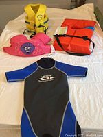 Lot items displayed on white fabric background including children's wetsuit, yellow adult life jacket, orange child life jacket and pink Puddle Jumper