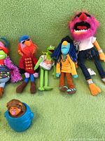 Plush dolls of Animal, Kermit in T-shirt, and Fraggle characters lying on green fabric background, and plastic Fraggle head in blue barrel included.