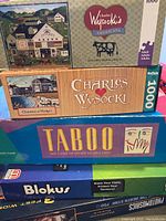 Stack of board games and puzzles including Charles Wysocki Americana puzzle, Taboo, Blokus, and a Photographics puzzle.