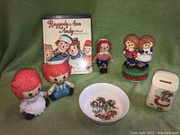 Full view of vintage Raggedy Ann and Andy dolls, porcelain bowl, music box, and booklet showing overall lot composition.
