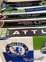 Six soccer scarves laid out showing various team logos and match dates including Seattle Sounders FC matches from 2010 and a Chelsea FC USA Tour 2012 scarf.