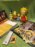 Photo of The Simpsons Clue game box, cards, game board, and stuffed character toys including Lisa, Bart, Maggie, and Marge Simpson.