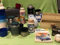 Photo showing vintage thermoses in various colors and sizes, including metal and plastic ones, along with a beige tackle box and golf ball boxes.
