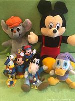 Wide shot showing all vintage Disney plush and plastic toys in the lot including Mickey Mouse, Donald Duck, Goofy, and others, placed against green fabric background.