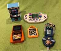 Five vintage handheld electronic games arranged on green surface, showing variety of game types and designs.