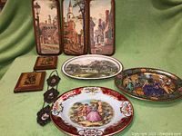 Wide shot of collection showing three rectangular Courtier & Ives tin platters with street scenes and one oval estate scene platter, plus Daher and detailed scene oval platters, and small framed plaques on decorative hanging