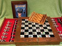 Photo shows large hand-carved chess set with wooden box open revealing red felt interior and hand carved chess pieces strapped inside, small chess set on top of box with lighter and darker wooden pieces arranged, Stratego game box upright behind the chess sets.