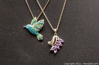 Two sterling silver necklaces with pendants on a black background. One pendant shaped like a hummingbird with blue and green rhinestones, the other resembling a branch with purple marquise-cut stones.