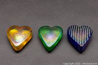 Front view of three iridescent heart-shaped art glass paperweights in amber, green, and blue with ribbed texture, displayed side by side.