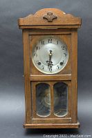 Full frontal image of the antique wooden wall clock showing carved shell decoration, clock face with Arabic numerals, and glass pendulum door.