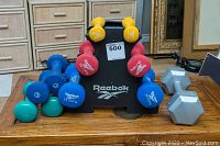 Overall photo of Reebok hand weights arranged on the rack and wooden table, includes various weight colors and grey hex dumbbells.