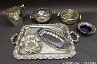 Various silver plated serving items arranged including trays, pitcher, covered dish, and decorative glass bowls.