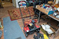 Full view of the red and black Toro Power Curve electric snow blower with orange extension cord, placed indoors on a wooden floor near various household items.