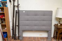 Gray upholstered headboard with horizontal tufts, minor wear on bottom edge, leaning against wall near metal bed frame parts and furniture.