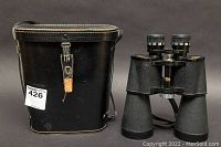 Binoculars standing upright next to their black leather carrying case showing exterior wear and a strap