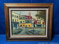 Framed oil painting depicting a colorful street scene with buildings and two pedestrians crossing a square, artist's signature visible.