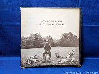 Front cover of George Harrison All Things Must Pass vinyl album showing a image of Harrison and children in an outdoor setting. Cover has some wear and small corner damage.