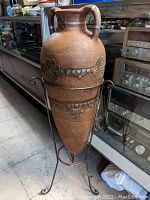 Front and side view of the large clay pot on a metal stand showing the texture and floral decorative elements.