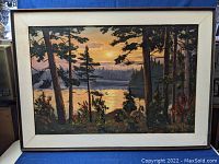 Full view of the large framed landscape painting depicting a lakeside scene through tall pine trees at sunset or sunrise