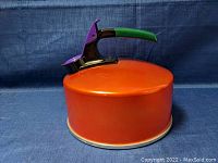Side view showing the rounded orange body with black, green and purple handle and spout lever