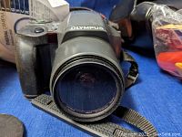 Front view of Olympus SLR camera with attached zoom lens, showing brand name on body and lens glass.