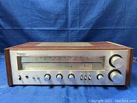 Front view of Technics SA-200 stereo receiver showing tuning dial, power switch, and various control knobs on a silver faceplate within a wood veneer cabinet.