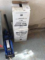 Two white boxes of MotoMaster Axle Stands stacked vertically beside a blue PowerFist floor jack on garage floor.